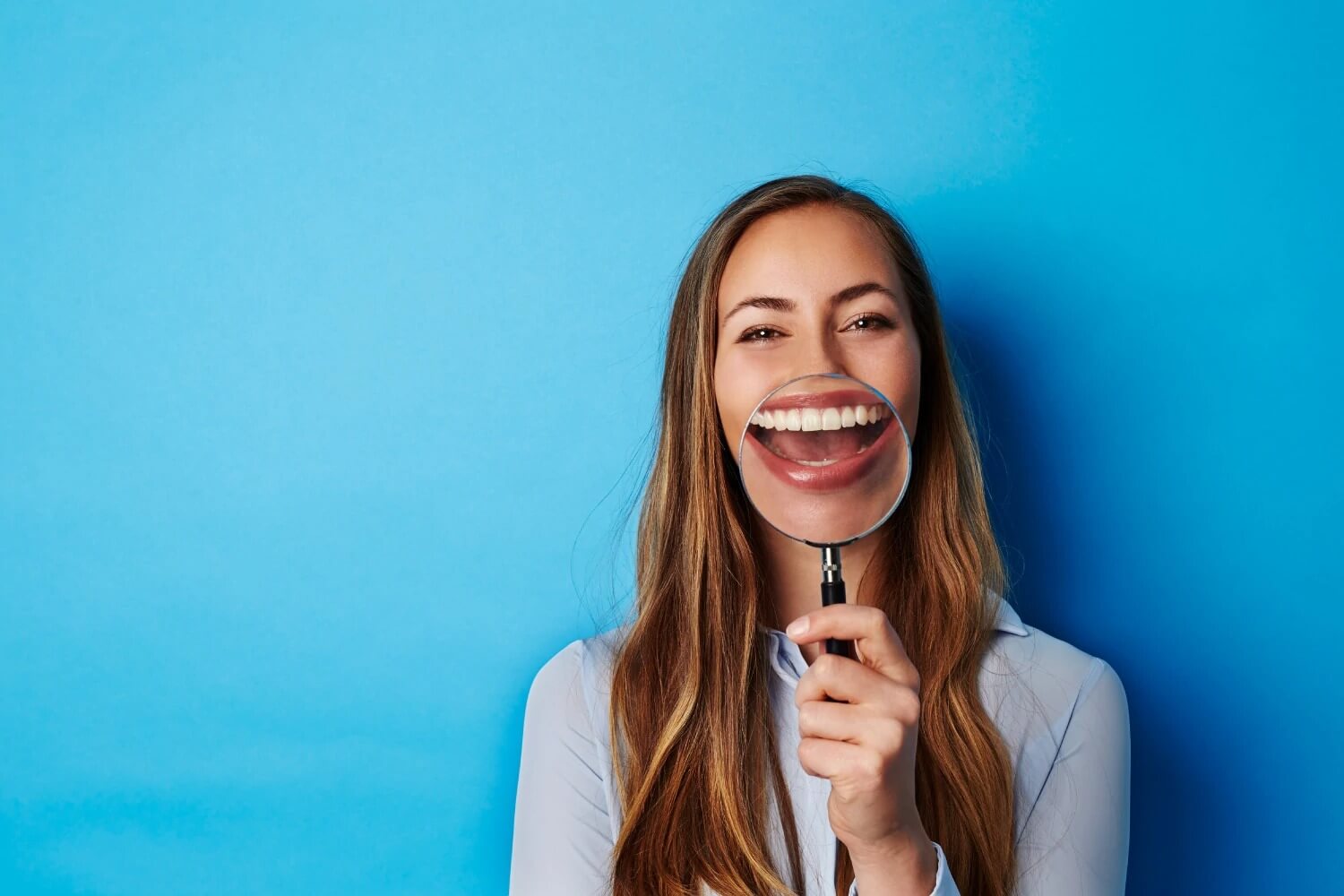 White brunette woman smiles as she holds up a magnifying glass to her teeth against a bright blue wall
