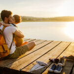 A dad hugs his son while sitting on a dock overlooking a lake as the sun sets