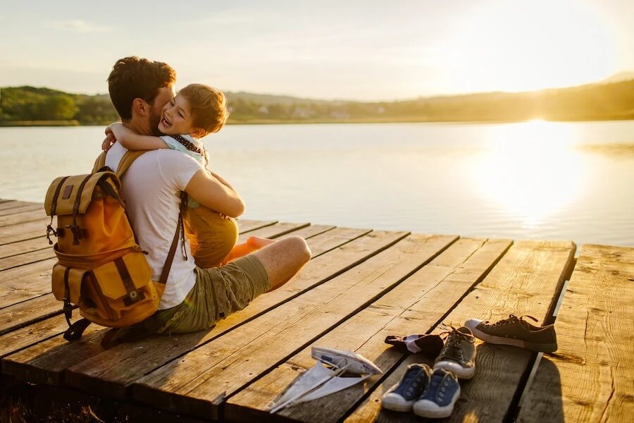 A dad hugs his son while sitting on a dock overlooking a lake as the sun sets