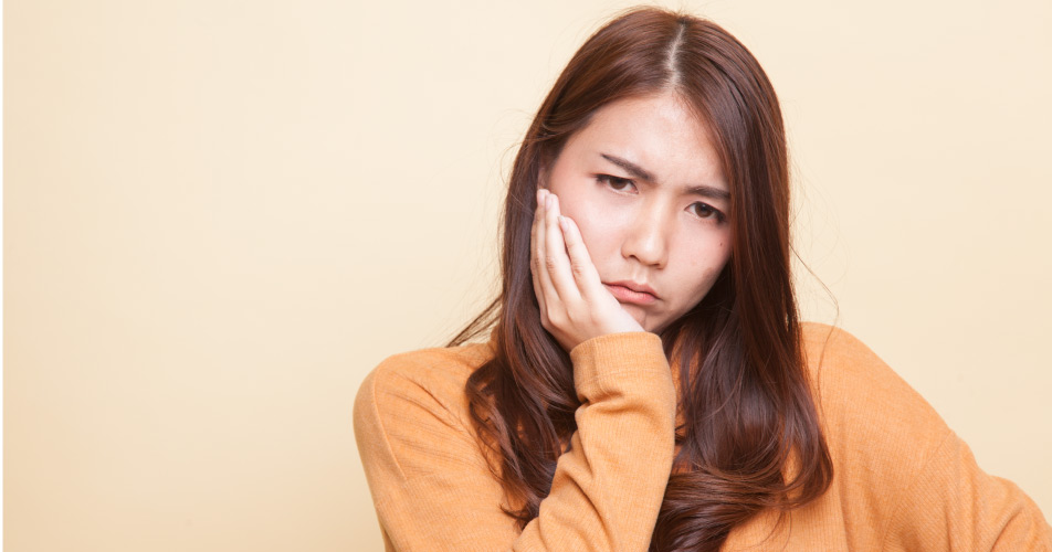 Dark-haired woman in a mango-colored shirt holds her jaw and cringes in pain because she needs root canal therapy