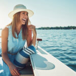 A brunette woman in a hat and sundress smiles while sitting on a boat on the water