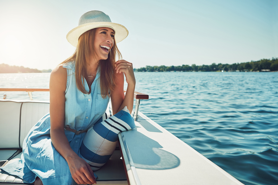 A brunette woman in a hat and sundress smiles while sitting on a boat on the water